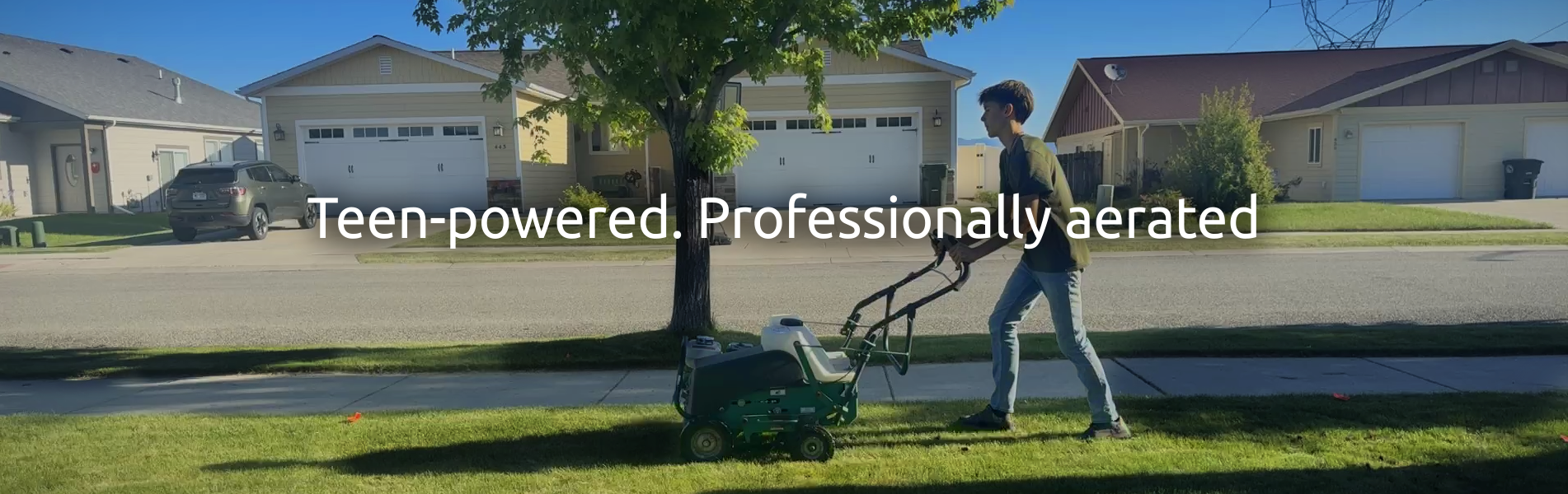 Flathead Aeration member pushing the aeration machine over green grass with houses behind and a view of Kalispell and Whitefish